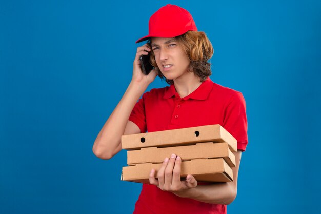 Jeune livreur suspect en uniforme rouge parlant au téléphone mobile tout en tenant des boîtes de pizza sur fond bleu isolé