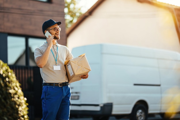 Photo gratuite jeune livreur tenant des colis tout en communiquant sur un téléphone portable et debout dans la rue