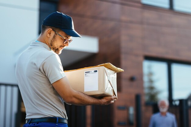 Jeune livreur transportant un colis et lisant l'adresse sur une étiquette Son client se tient en arrière-plan