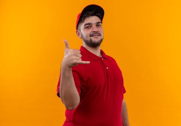 Jeune livreur en uniforme rouge et cap faisant appelez-moi geste avec sourire sur le visage