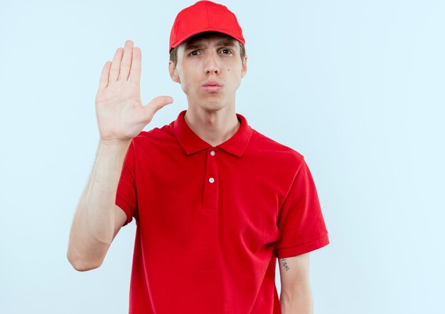 Jeune livreur en uniforme rouge et cap faisant panneau d'arrêt avec la main ouverte à l'avant avec un visage sérieux debout sur un mur blanc