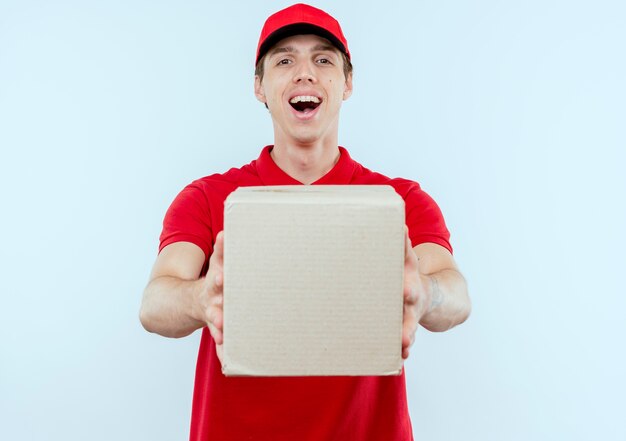 Jeune livreur en uniforme rouge et cap holding boîte en carton souriant joyeusement heureux et positif debout sur mur blanc