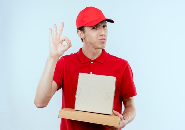 Jeune livreur en uniforme rouge et cap holding box package et boîte à pizza à l'avant souriant montrant signe ok debout sur mur blanc