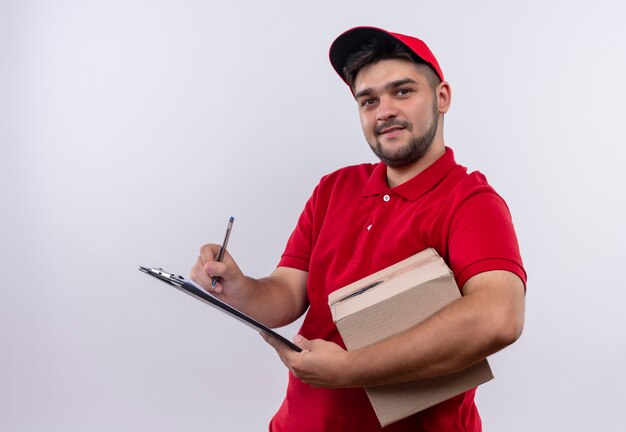 Jeune livreur en uniforme rouge et cap holding box package écrit sur une page vierge souriant sympathique