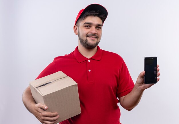 Jeune livreur en uniforme rouge et cap holding box package montrant smartphone regardant la caméra avec un sourire confiant