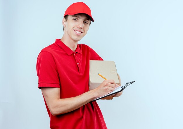 Jeune livreur en uniforme rouge et cap holding box package et presse-papiers écrit quelque chose de souriant à l'avant debout sur un mur blanc