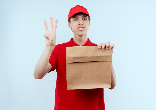 Jeune livreur en uniforme rouge et cap holding paper package montrant le numéro trois à la confiance debout sur mur blanc