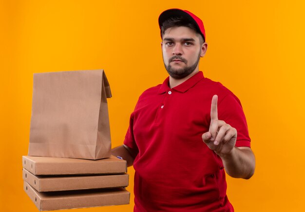 Jeune livreur en uniforme rouge et cap holding paper package et pile de boîtes de pizza montrant l'index d'avertissement avec visage sérieux