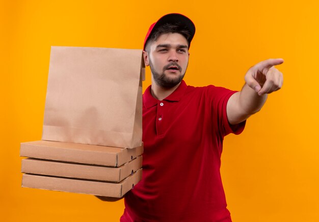 Jeune livreur en uniforme rouge et cap holding paper package et pile de boîtes à pizza pointant vers quelque chose avec une expression sceptique