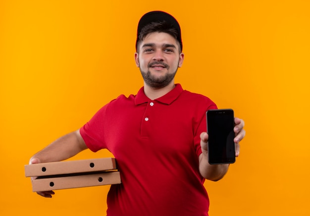 Jeune livreur en uniforme rouge et cap holding pile de boîtes à pizza montrant smartphone souriant sympathique
