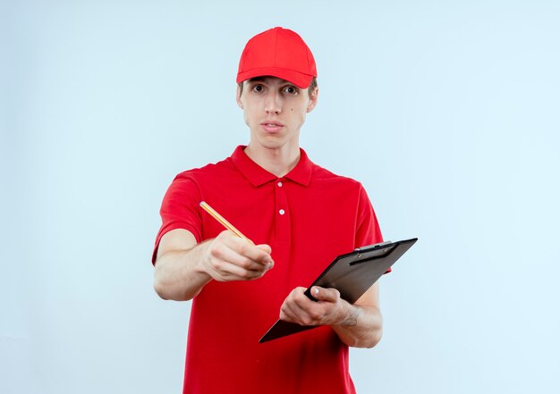 Jeune livreur en uniforme rouge et cap holding presse-papiers et crayon à l'avant avec le bras comme demandant debout sur un mur blanc