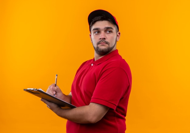 Jeune livreur en uniforme rouge et cap holding presse-papiers avec stylo à côté avec visage sérieux