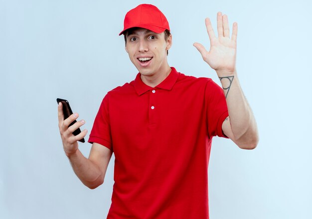 Jeune livreur en uniforme rouge et cap holding smartphone regardant vers l'avant en agitant avec la main debout sur un mur blanc