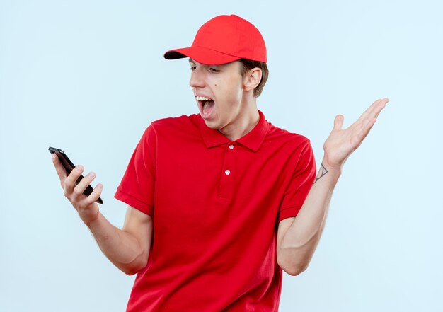 Jeune livreur en uniforme rouge et cap holding smartphone à surpris et confus avec bras levé debout sur un mur blanc