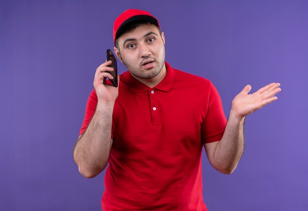 Jeune livreur en uniforme rouge et cap parler sur téléphone mobile à la confusion en haussant les épaules debout sur le mur violet