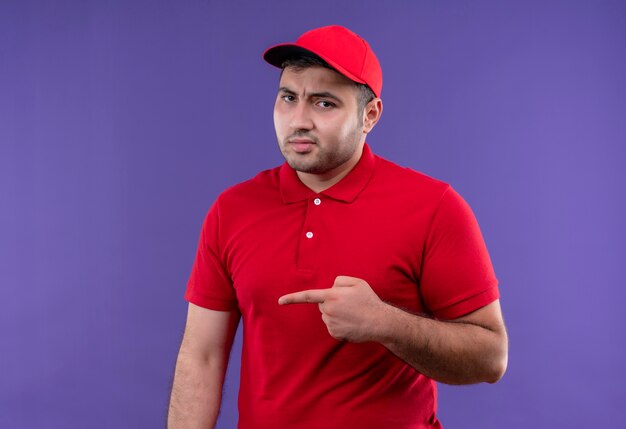 Jeune livreur en uniforme rouge et cap pointant avec le doigt sur le côté mécontent de se tenir debout sur un mur violet