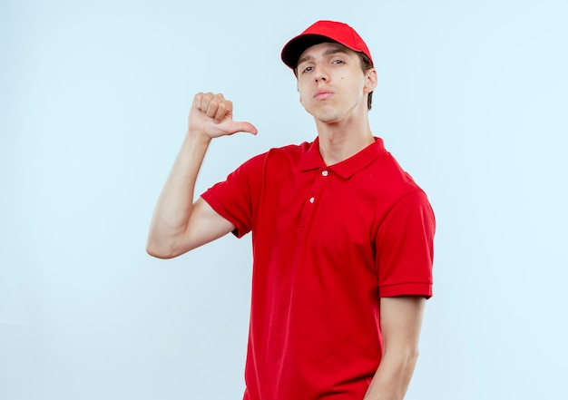 Jeune livreur en uniforme rouge et cap pointant avec le doigt sur lui-même à la confiance de soi-même debout sur un mur blanc
