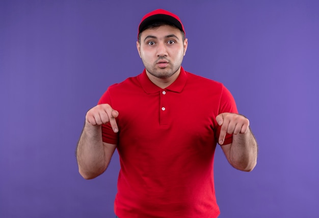 Jeune livreur en uniforme rouge et cap pointant avec l'index vers le bas à la surprise debout sur le mur violet