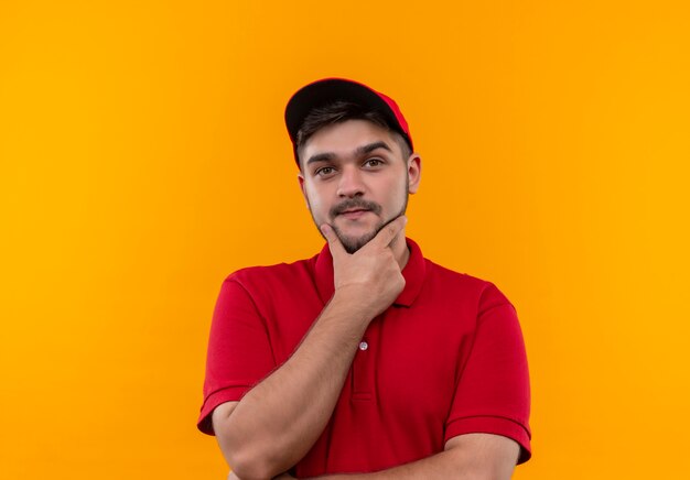 Jeune livreur en uniforme rouge et cap regardant la caméra avec la main sur le menton avec une expression confiante sérieuse