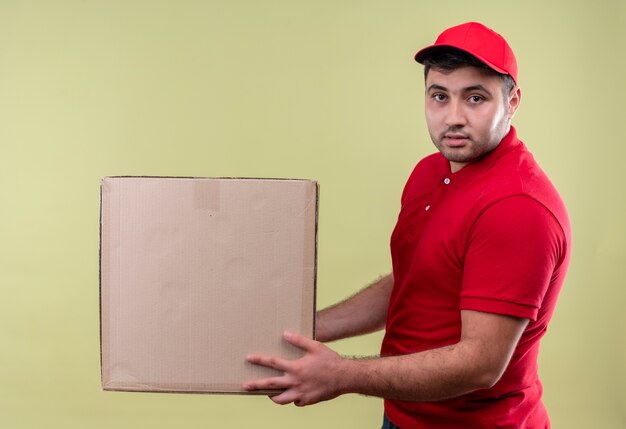 Jeune livreur en uniforme rouge et cap tenant une boîte en carton avec une expression confiante debout sur un mur vert