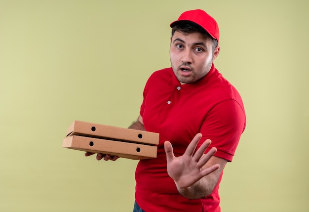 Photo gratuite jeune livreur en uniforme rouge et cap tenant des boîtes de pizza faisant panneau d'arrêt avec la main avec l'expression de la peur debout sur le mur vert