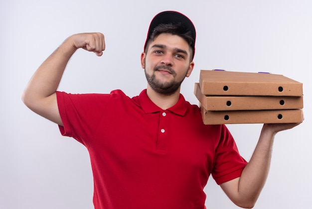 Jeune livreur en uniforme rouge et cap tenant des boîtes à pizza levant le poing montrant les biceps, concept gagnant