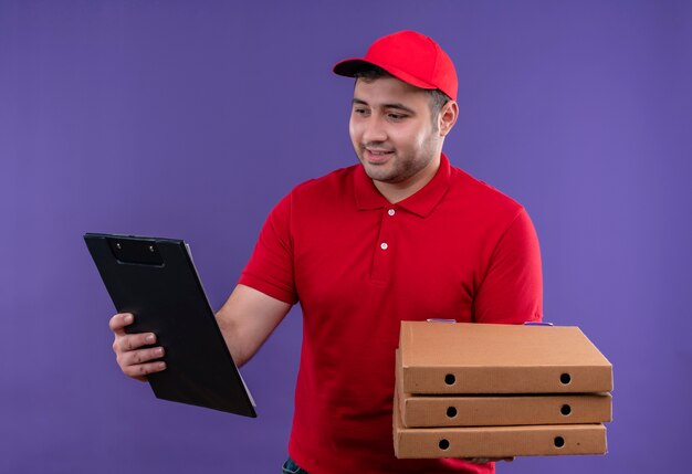 Jeune livreur en uniforme rouge et cap tenant des boîtes à pizza et presse-papiers souriant avec visage heureux debout sur le mur violet