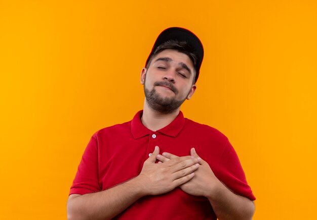 Jeune livreur en uniforme rouge et cap tenant les mains croisées sur la poitrine avec les yeux fermés ressentir des émotions positives