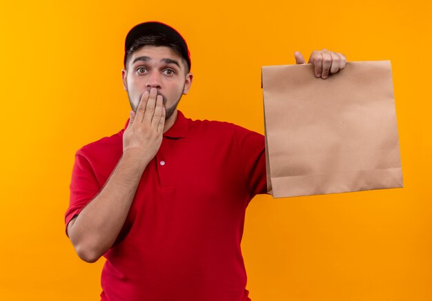 Jeune livreur en uniforme rouge et cap tenant le paquet de papier à la bouche surprise couvrant avec la main