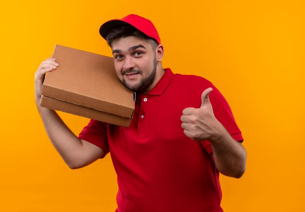 Jeune livreur en uniforme rouge et cap tenant la pile de boîtes à pizza montrant les pouces vers le haut en souriant