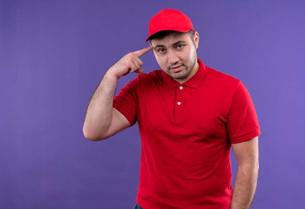 Photo gratuite jeune livreur en uniforme rouge et capuchon pointant avec le doigt son temple à la confiance debout sur le mur violet