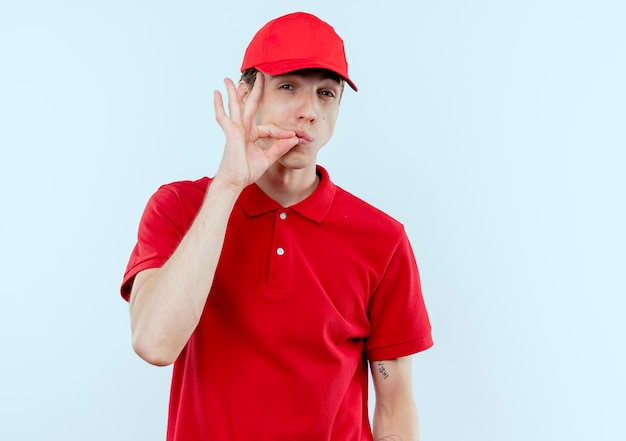 Jeune livreur en uniforme rouge et casquette faisant un geste de silence en fermant sa bouche avec une fermeture éclair debout sur un mur blanc