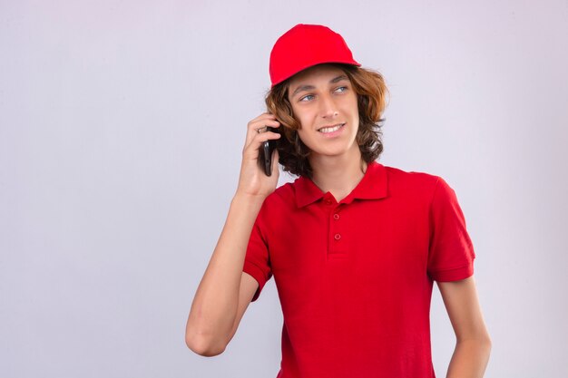 Jeune livreur en uniforme rouge parlant au téléphone mobile à côté avec sourire sur le visage sur fond blanc isolé