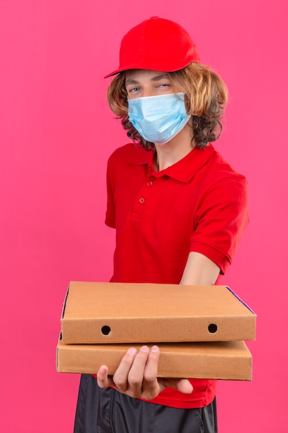 Jeune livreur en uniforme rouge portant un masque médical s'étendant sur des boîtes de pizza smiling friendly debout sur fond rose isolé