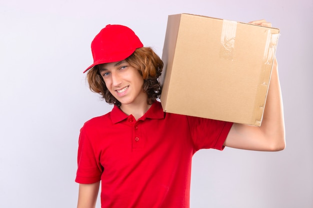 Jeune livreur en uniforme rouge tenant une boîte en carton sur l'épaule en regardant la caméra souriant sympathique sur fond blanc isolé