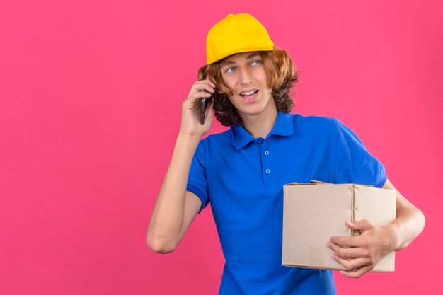 Jeune livreur vêtu d'un polo bleu et une casquette jaune tenant une boîte en carton tout en parlant sur smartphone avec souriant sympathique debout sur fond rose isolé