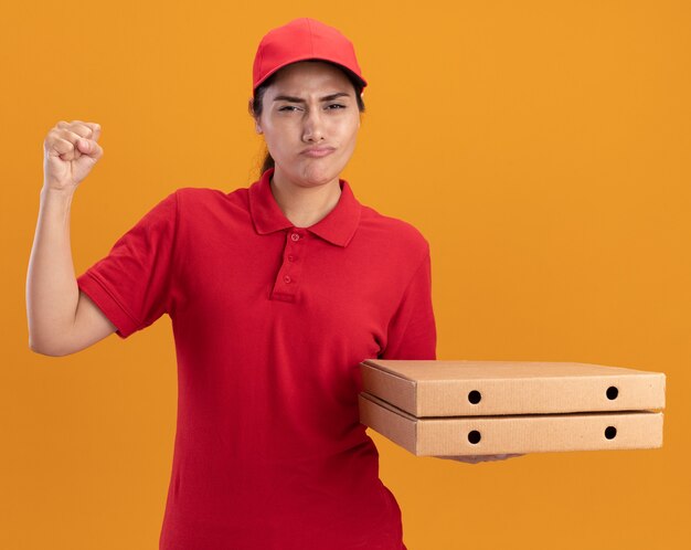 Jeune livreuse confiante portant un uniforme et une casquette tenant des boîtes à pizza levant le poing isolé sur un mur orange