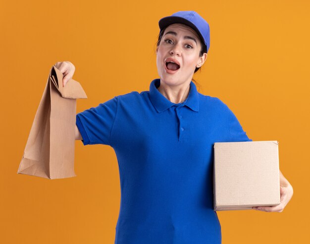 Jeune livreuse impressionnée en uniforme et casquette tenant une boîte à cartes et un paquet de papier isolé sur un mur orange