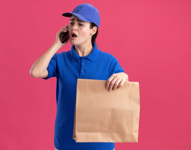 Jeune livreuse inquiète en uniforme et casquette tenant un paquet de papier parlant au téléphone regardant vers le bas isolé sur un mur rose avec espace de copie