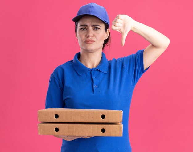 Jeune livreuse mécontente en uniforme et casquette tenant des paquets de pizza montrant le pouce vers le bas