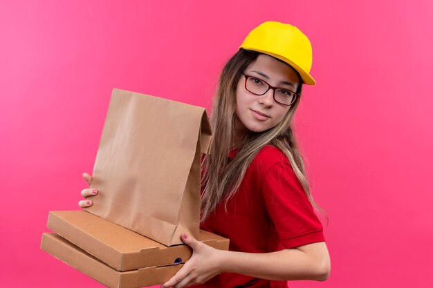 Jeune livreuse en polo rouge et casquette jaune tenant des boîtes à pizza et paquet de papier à la confiance