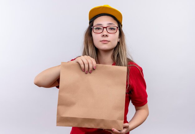 Jeune livreuse en polo rouge et casquette jaune tenant un paquet de papier regardant la caméra avec une expression confiante sérieuse