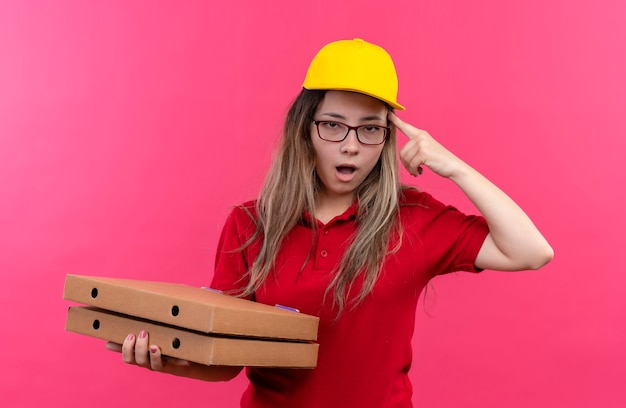 Jeune livreuse en polo rouge et casquette jaune tenant pile de boîtes de pizza pointant son temple, oublié, souvenez-vous de l'erreur, mauvaise mémoire concept