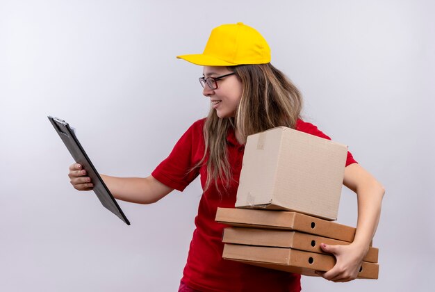 Jeune livreuse en polo rouge et casquette jaune tenant pile de boîtes de pizza à la recherche de presse-papiers dans son autre main à la confusion