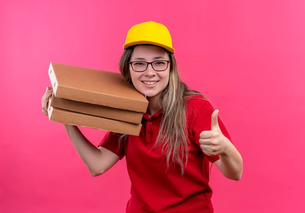 Jeune livreuse en polo rouge et casquette jaune tenant pile de boîtes à pizza regardant la caméra en souriant joyeusement montrant les pouces vers le haut