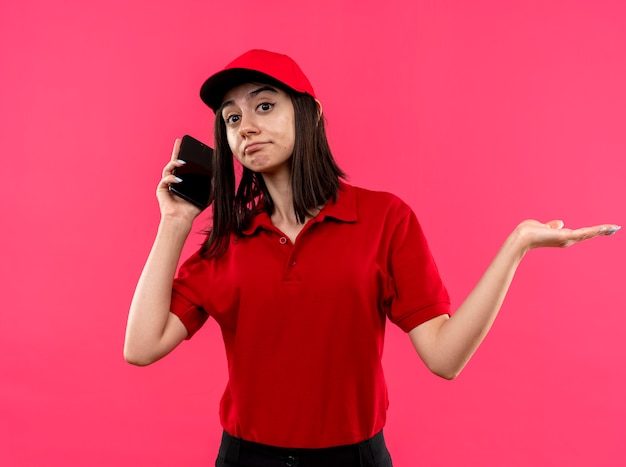 Jeune livreuse portant un polo rouge et une casquette à bras écarté confus sur le côté tout en parlant au téléphone mobile debout sur fond rose