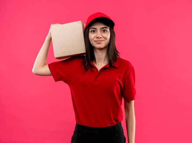 Jeune livreuse portant un polo rouge et une casquette tenant le paquet sur l'épaule avec le sourire sur le visage debout sur le mur rose