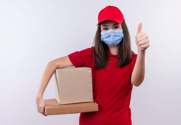 Jeune livreuse portant un t-shirt rouge en bonnet rouge porte un masque tenant une boîte et une boîte à pizza montrant les pouces vers le haut sur fond blanc isolé