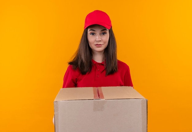 Jeune livreuse portant un t-shirt rouge en bonnet rouge tenant une grande boîte sur fond jaune isolé