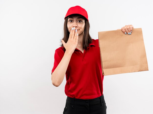 Jeune livreuse portant l'uniforme rouge et cap holding paper package looking at camera surpris coneting bouche avec main debout sur fond blanc